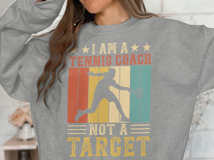 Gray sweatshirt with graphic design of a tennis player and text "I am a tennis coach not a target".