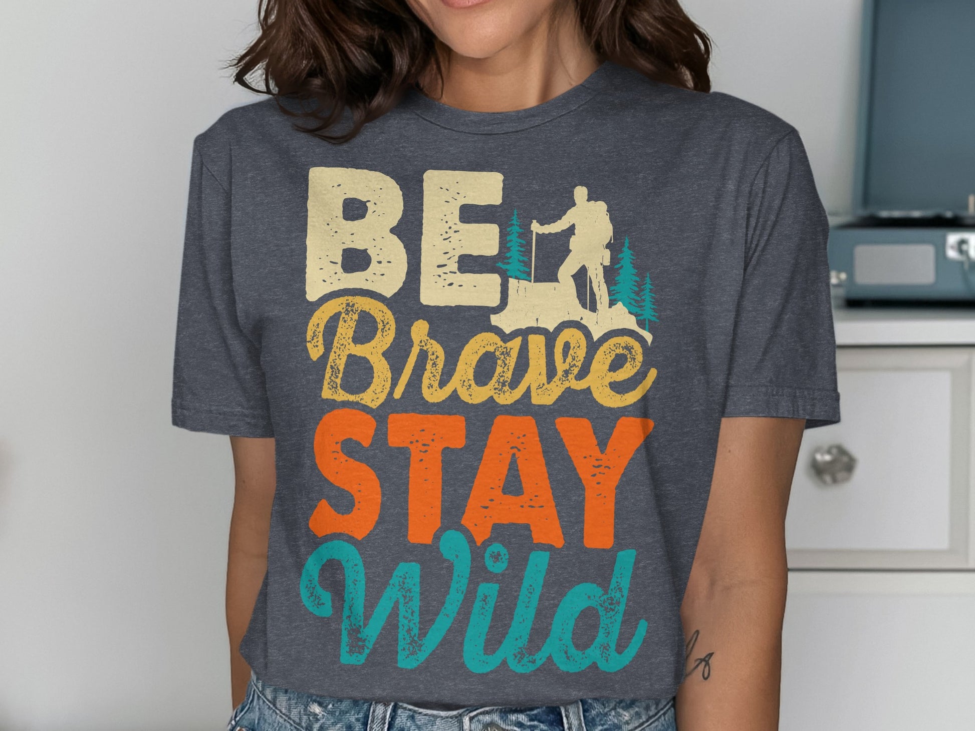 BE Brave STAY Wild text with hiker and mountain graphic on a charcoal t-shirt.