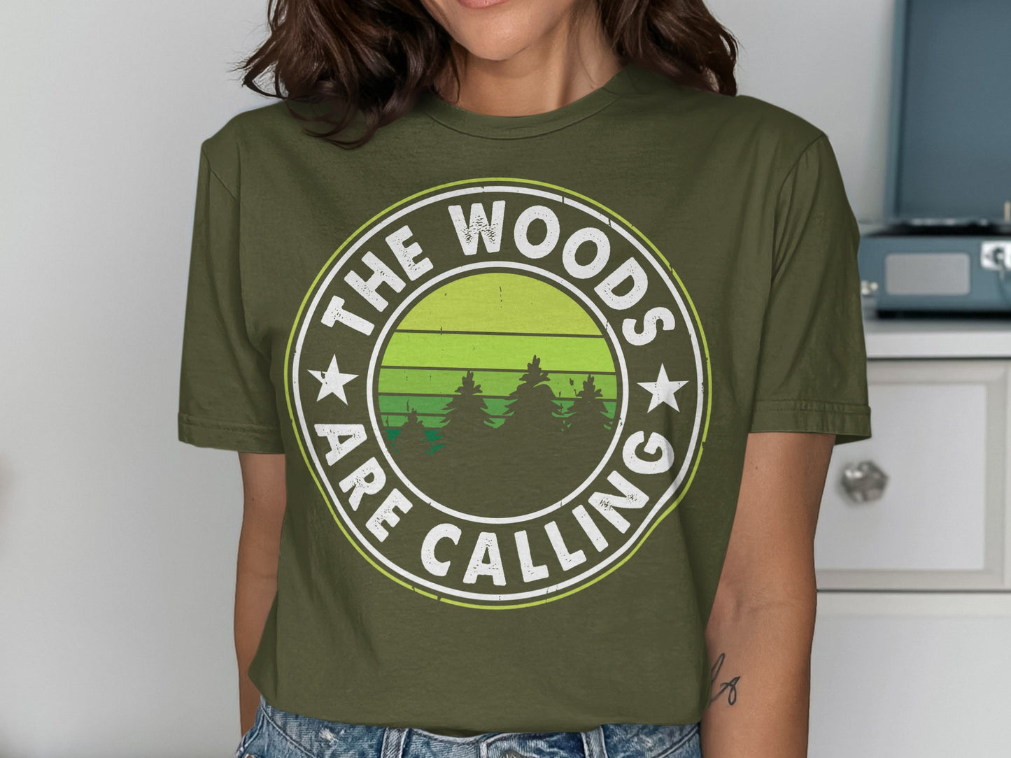 Olive green t-shirt with "The Woods Are Calling" text and forest silhouette.