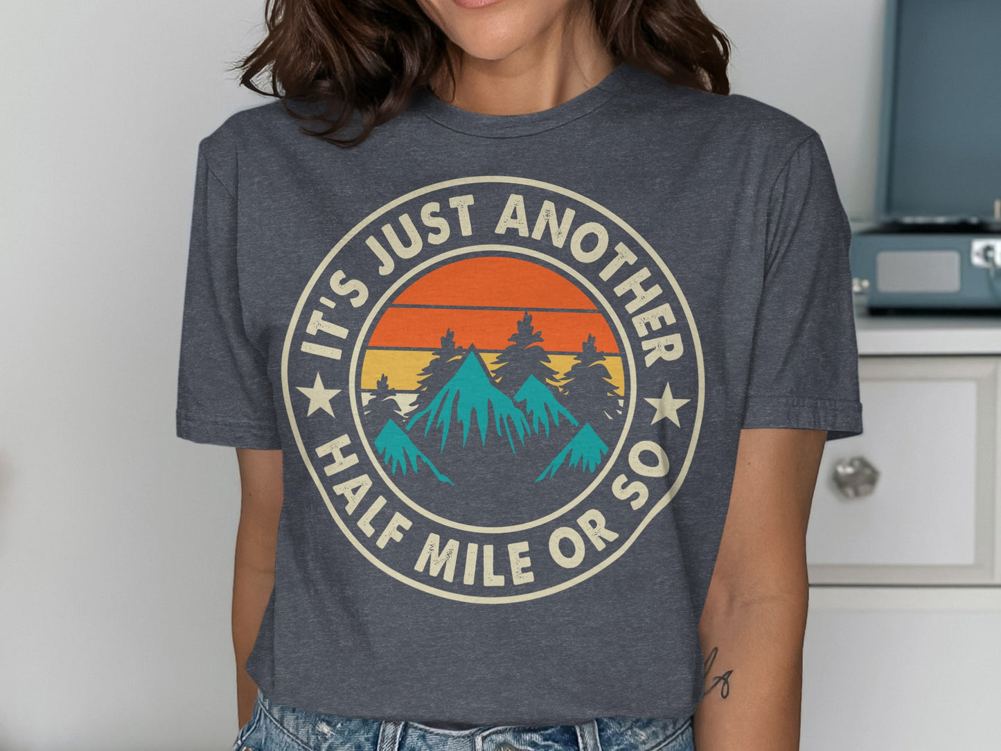Circular graphic with mountains, trees, sun, and text: "IT'S JUST ANOTHER HALF MILE OR SO".