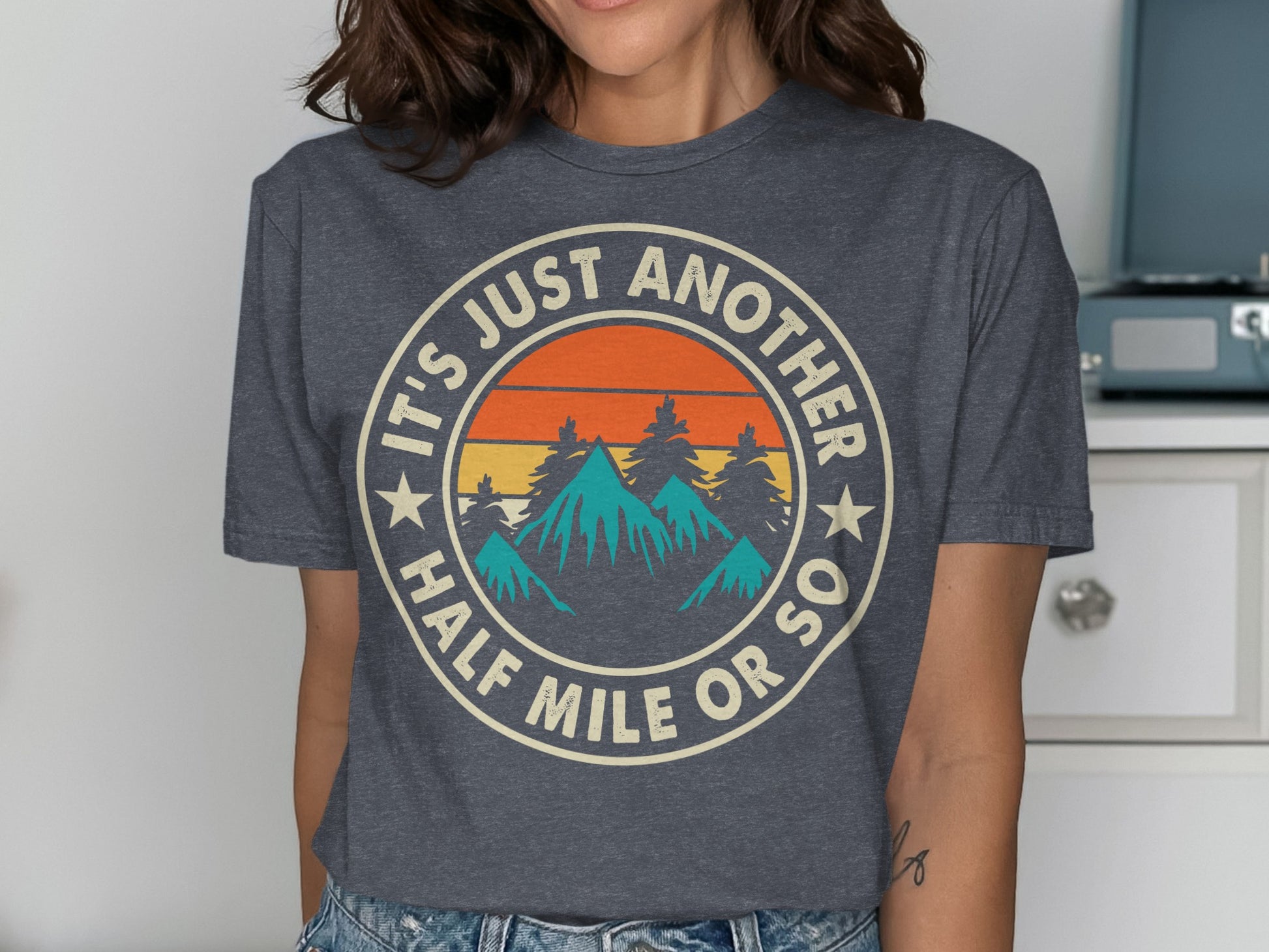 Circular graphic with mountains, trees, sun, and text: "IT'S JUST ANOTHER HALF MILE OR SO".