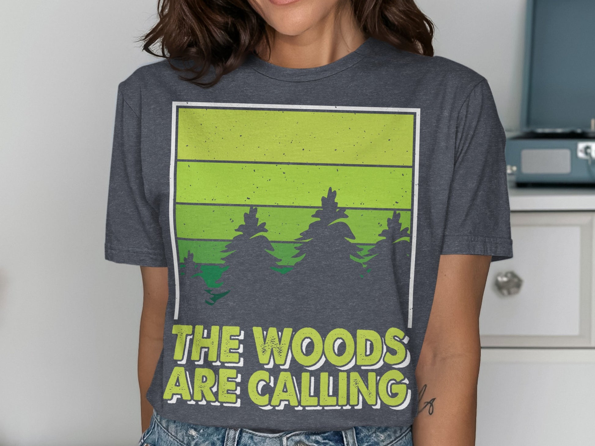 Dark grey t-shirt with green striped forest scene and text "The Woods Are Calling".
