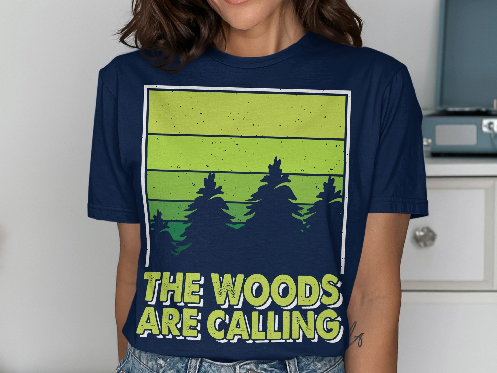 Dark blue t-shirt with green striped forest graphic and text "THE WOODS ARE CALLING".