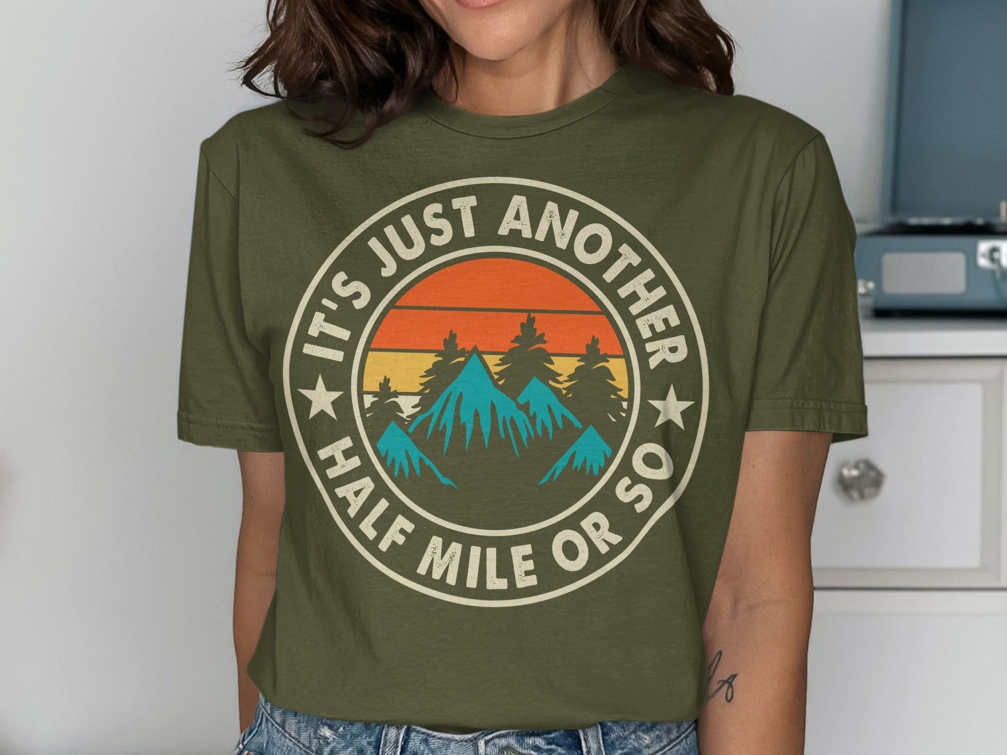 Green t-shirt with circular graphic: mountains, trees, sunset, and text "IT'S JUST ANOTHER HALF MILE OR SO".