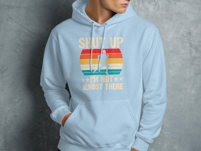 Light blue hoodie with text "SHUT UP I'M NOT *** ALMOST THERE" and retro sunset graphic.
