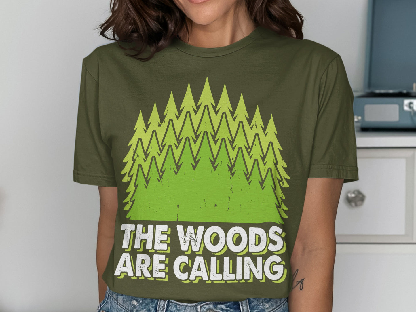 Green t-shirt with stylized pine trees and "The Woods Are Calling" text.