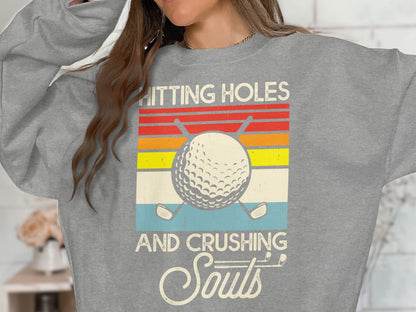Grey sweatshirt with a retro graphic of a golf ball and clubs, text reads "HITTING HOLES AND CRUSHING SOULS".