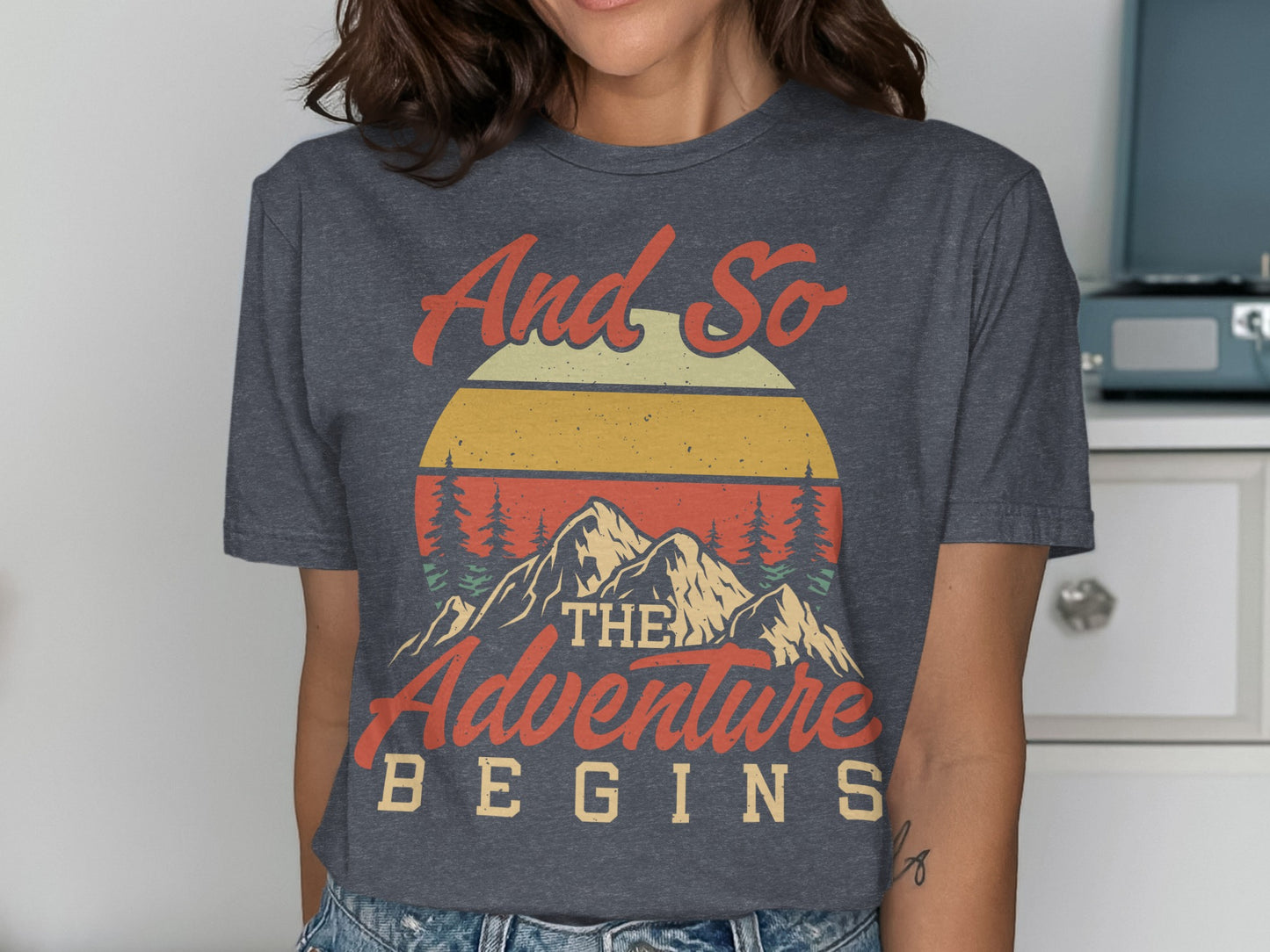 Dark grey t-shirt with sunset, mountains, and text "And So The Adventure Begins".