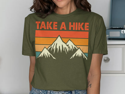 Olive green t-shirt with "TAKE A HIKE" in retro font above snowy mountain graphic.
