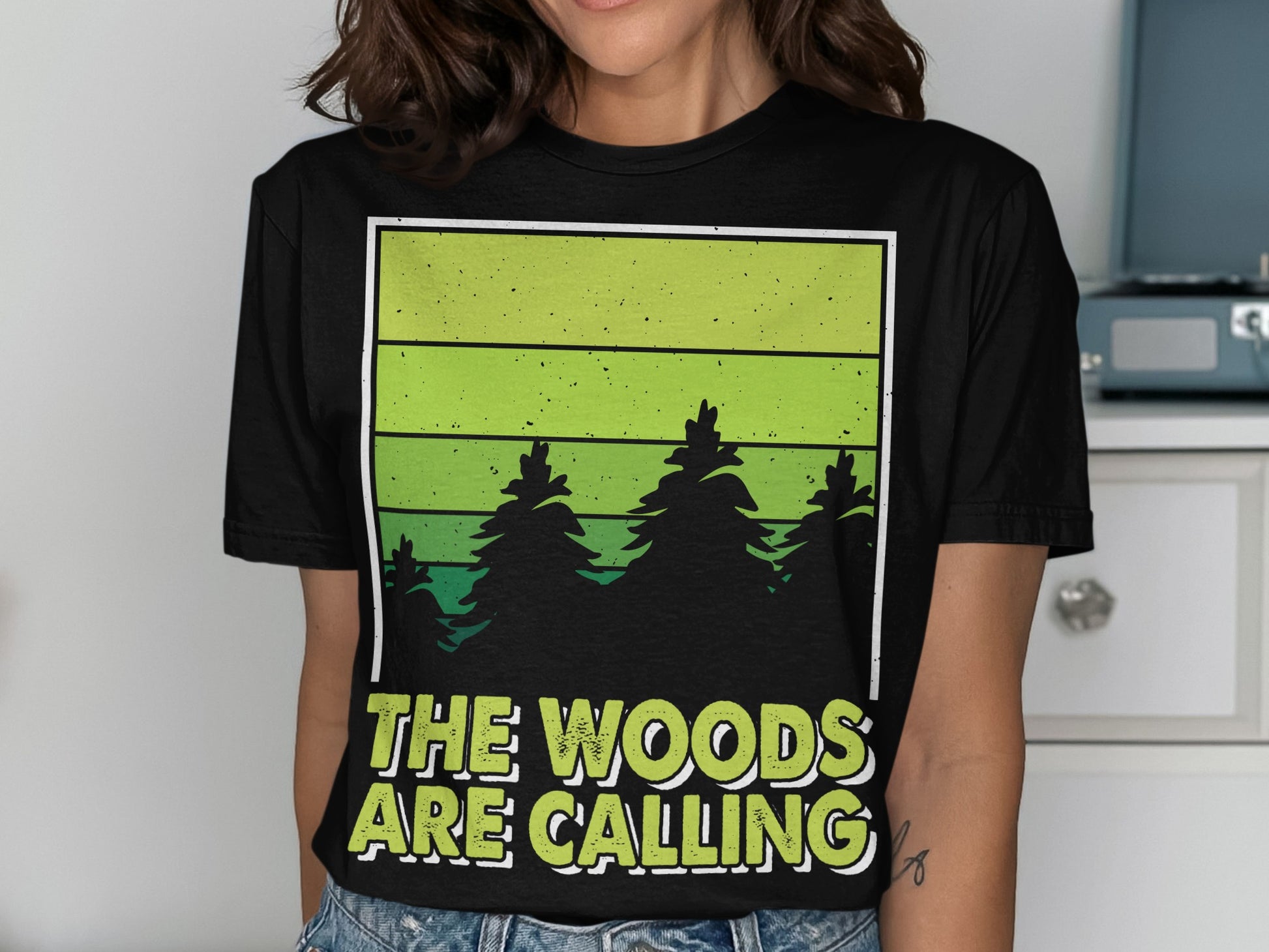 Black t-shirt with green gradient and pine trees, text "THE WOODS ARE CALLING".