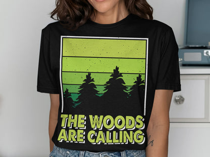 Black t-shirt with green gradient and pine trees, text "THE WOODS ARE CALLING".