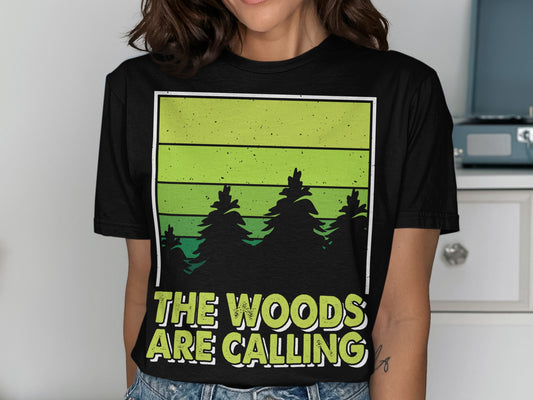 Black t-shirt with green gradient and pine trees, text "THE WOODS ARE CALLING".