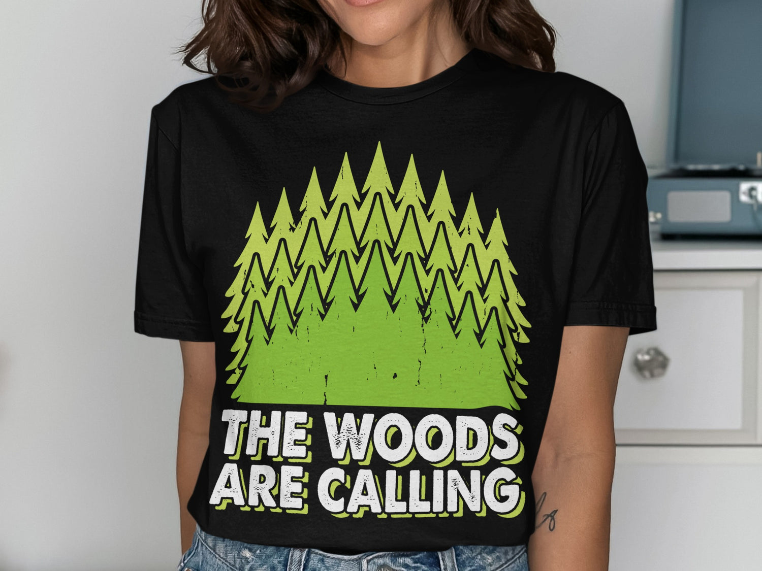 Black t-shirt with green pine trees and text "THE WOODS ARE CALLING".