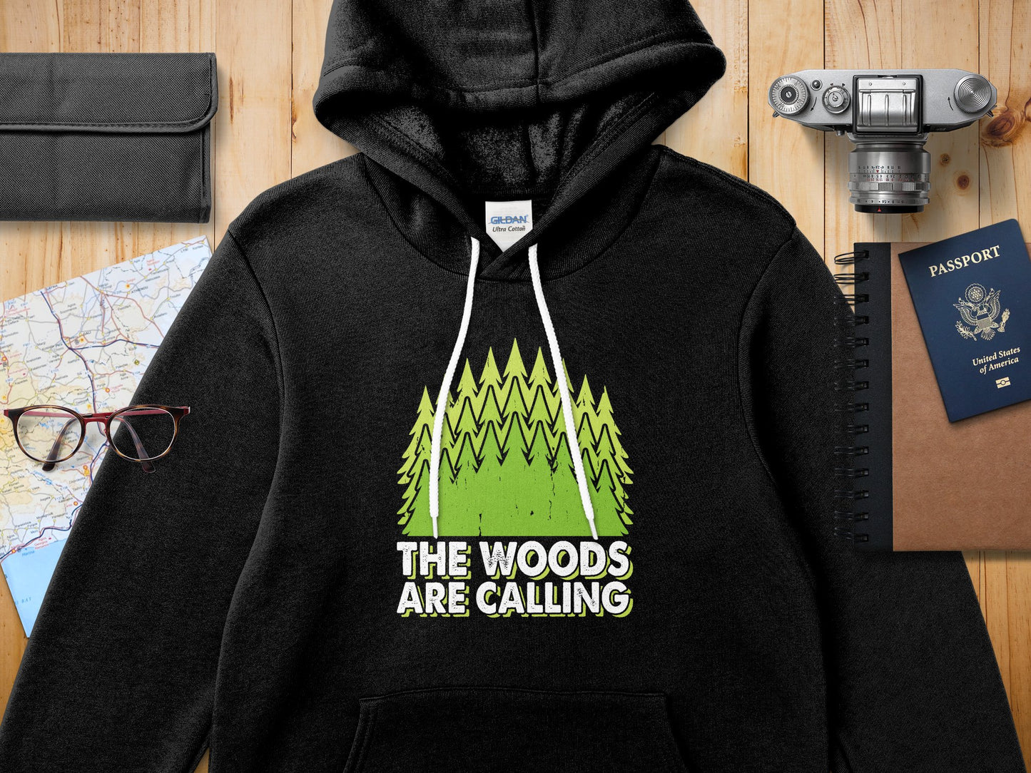 Black Gildan hoodie with green trees and "The Woods Are Calling" text.