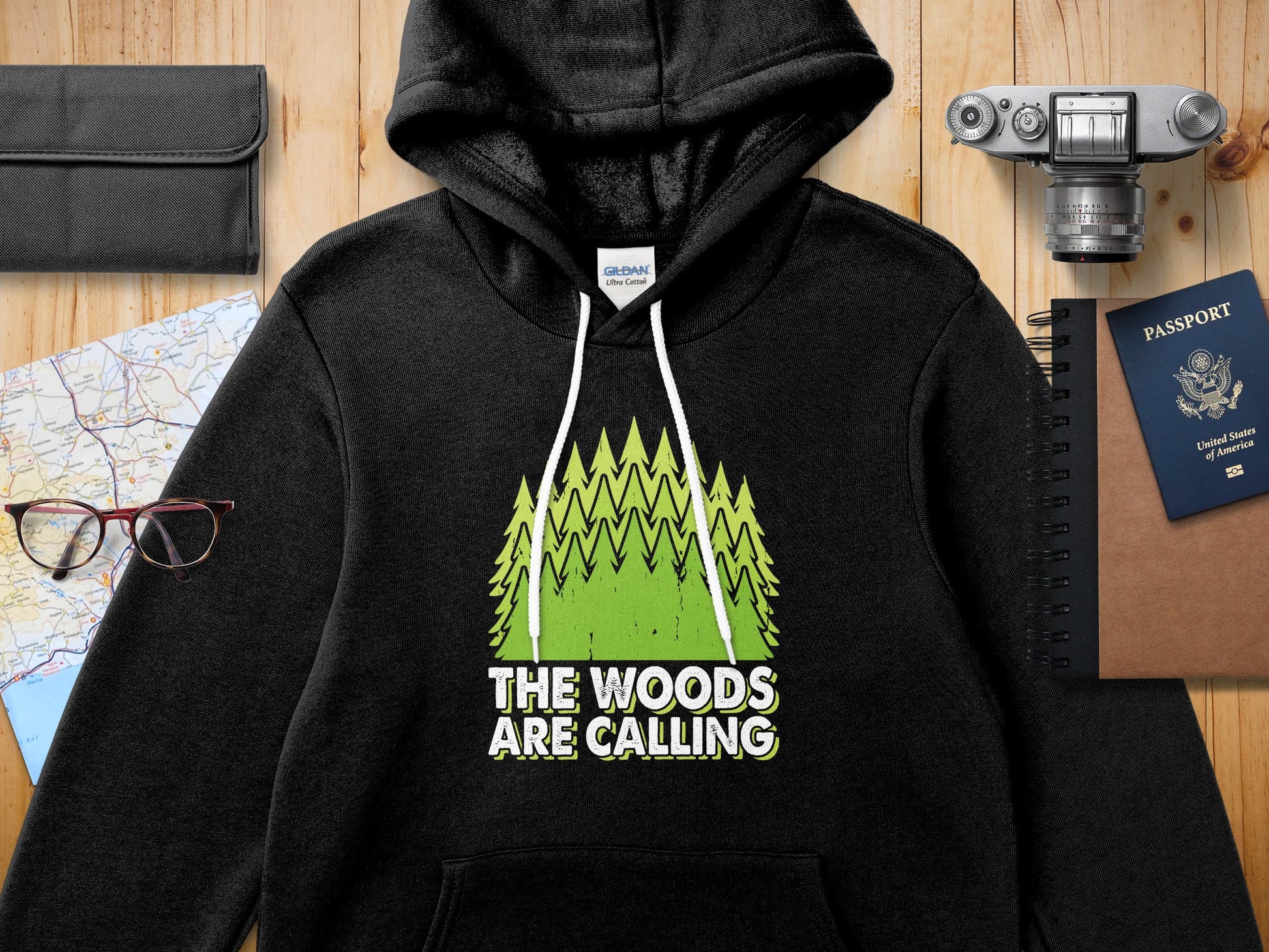Black Gildan hoodie with green trees and "The Woods Are Calling" text.