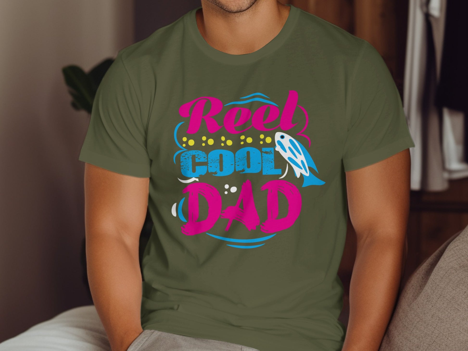 "Reel Cool Dad" text with fishing lure graphic on olive green t-shirt.