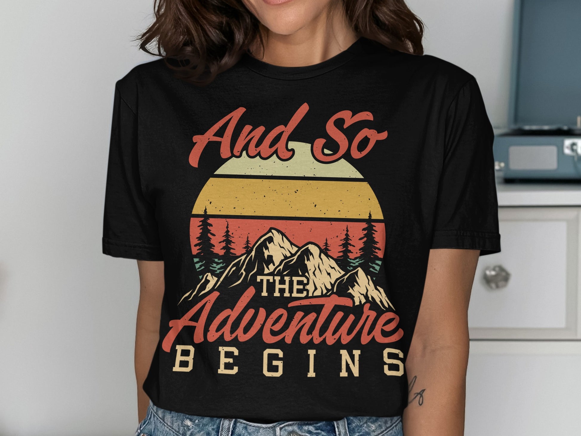 Black t-shirt with mountains, sunset, and text "And so the adventure begins".