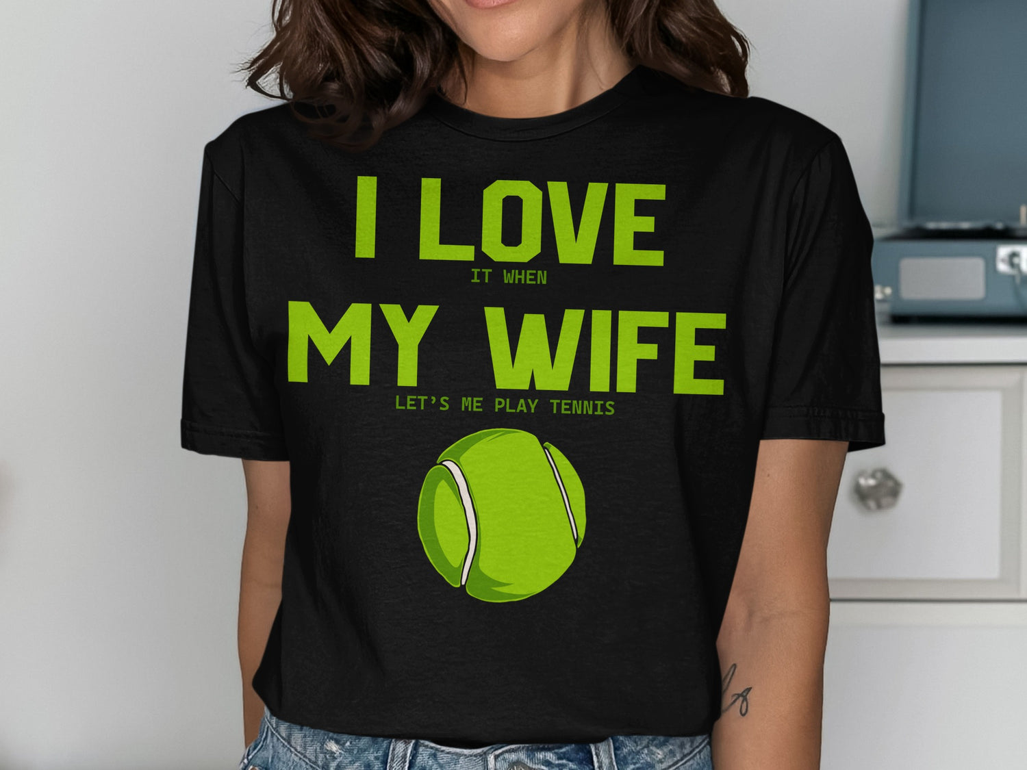 Black t-shirt with green text reading "I LOVE IT WHEN MY WIFE LETS ME PLAY TENNIS" and a tennis ball graphic.