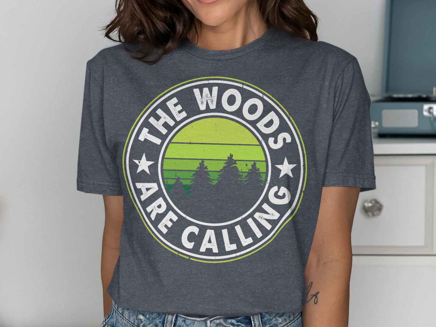Dark grey t-shirt with white text "The Woods Are Calling" and green forest graphic.