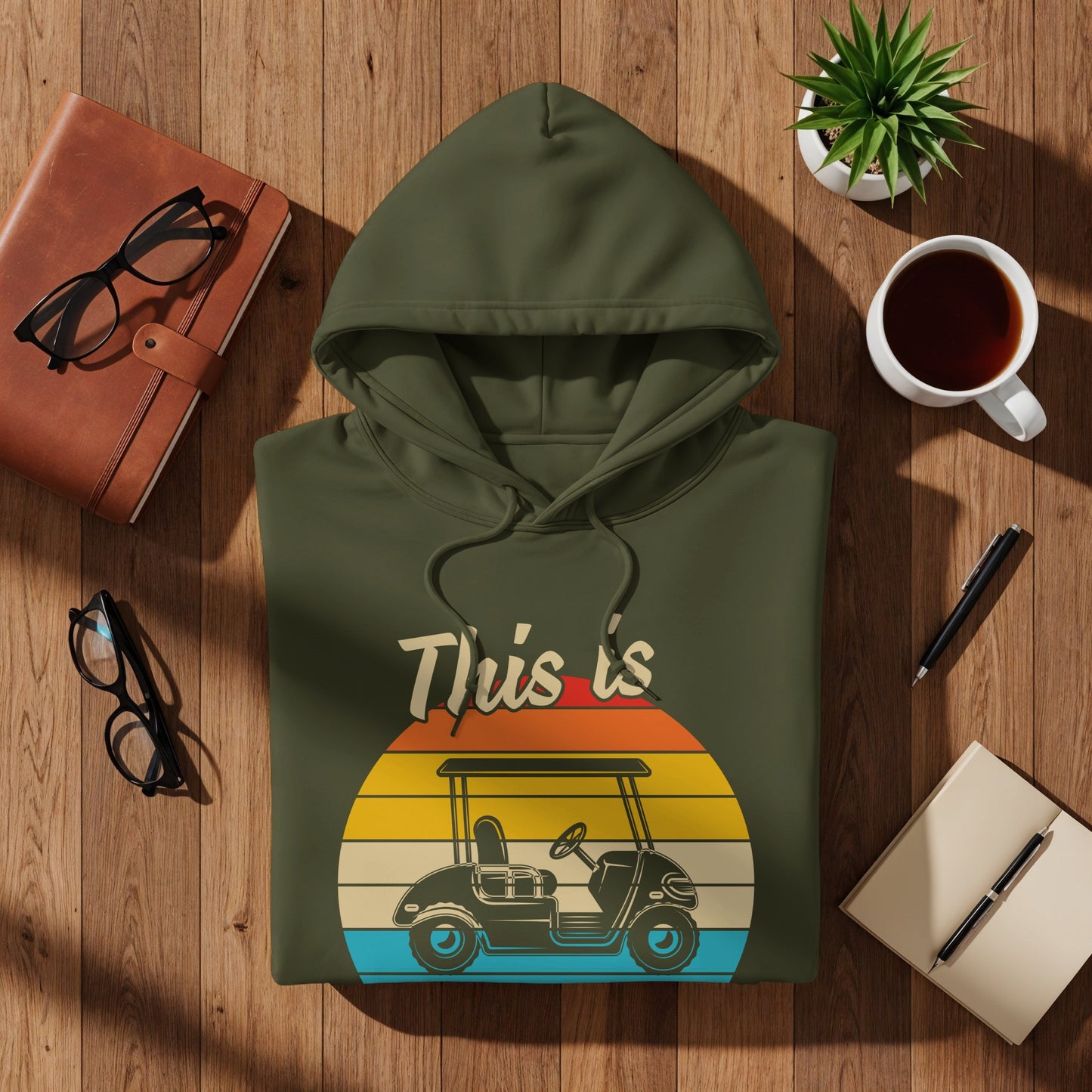 Green hoodie with "This is" text and golf cart graphic on a sunset background.