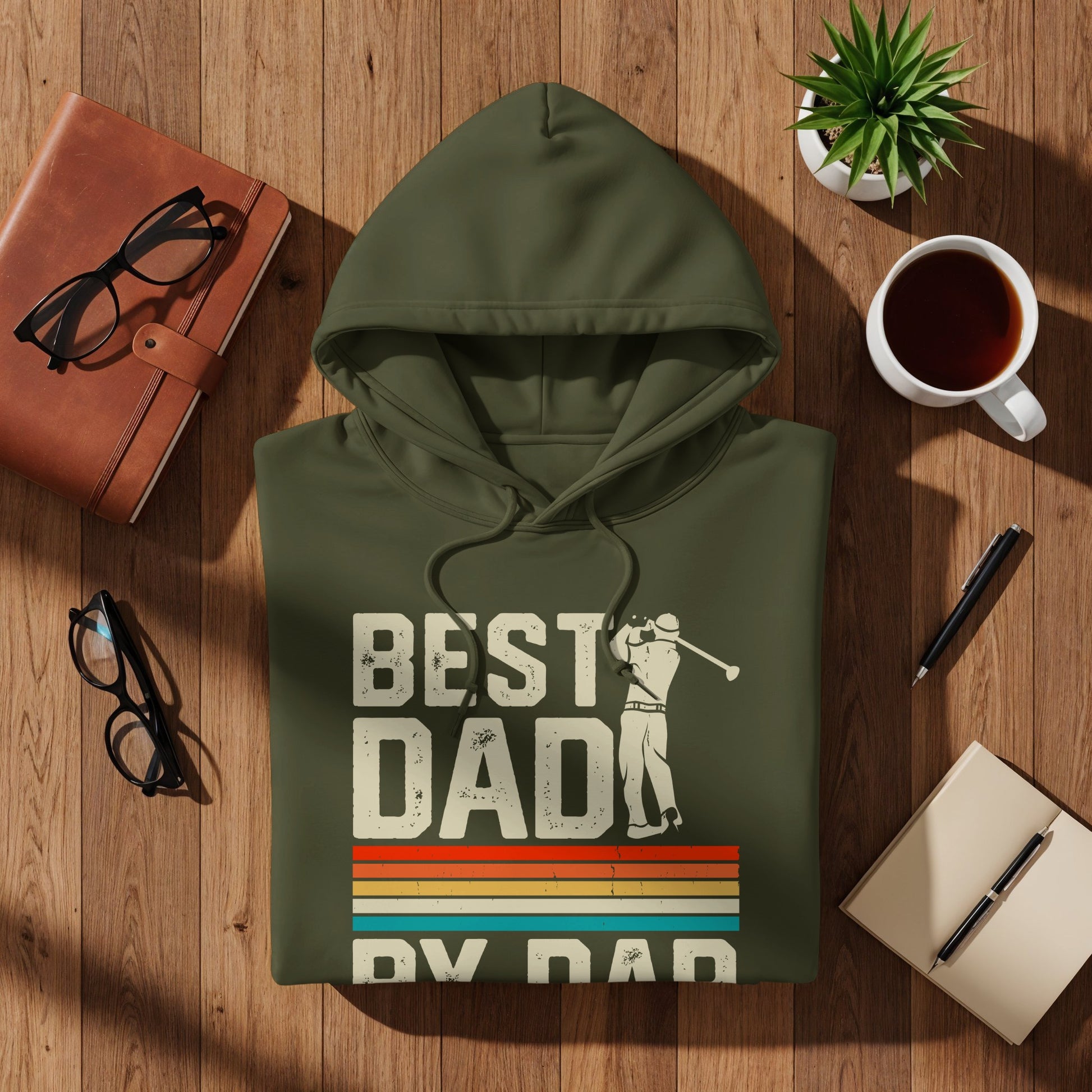 Green hoodie with "Best Dad" and golf design, flat lay on wood.