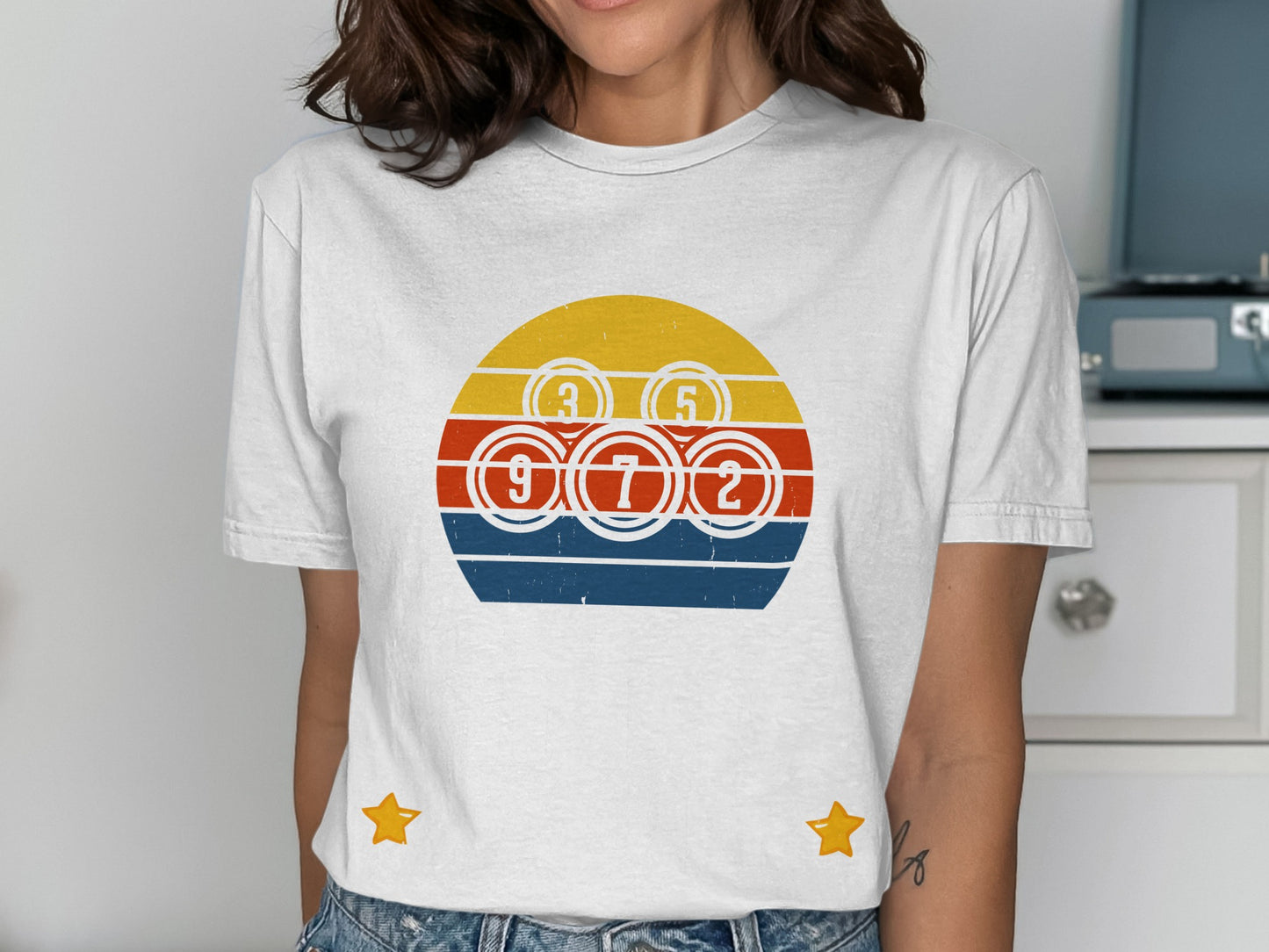 White t-shirt with a circular design featuring bingo balls and a retro sunset graphic.