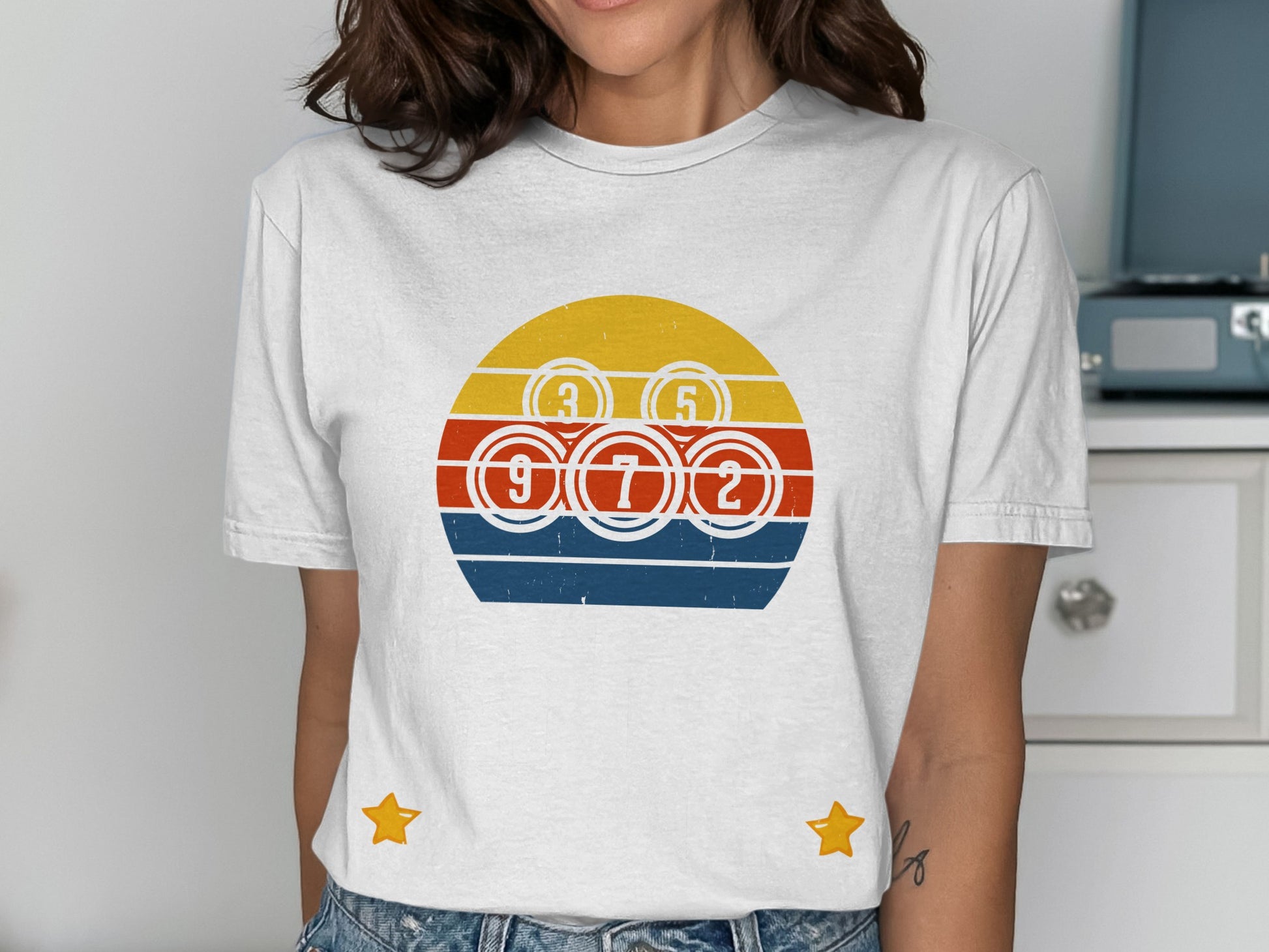 White t-shirt with a circular design featuring bingo balls and a retro sunset graphic.