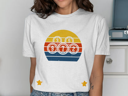 White t-shirt with a circular design featuring bingo balls and a retro sunset graphic.