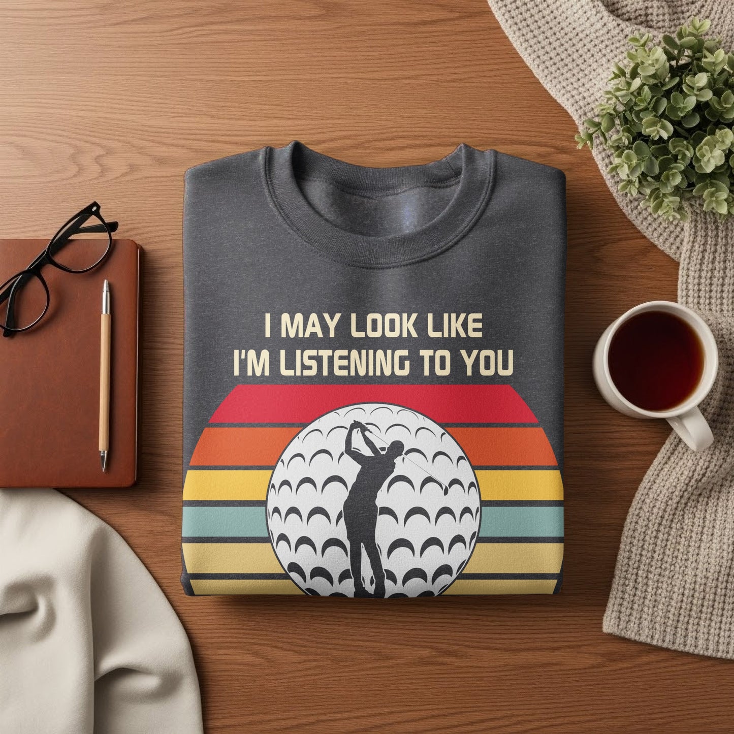 Dark sweatshirt with a golf ball and sunset graphic, text reads "I MAY LOOK LIKE I'M LISTENING TO YOU".