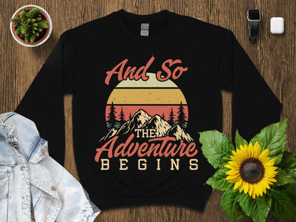 Black sweatshirt with "And So The Adventure Begins" graphic and mountain sunset design.