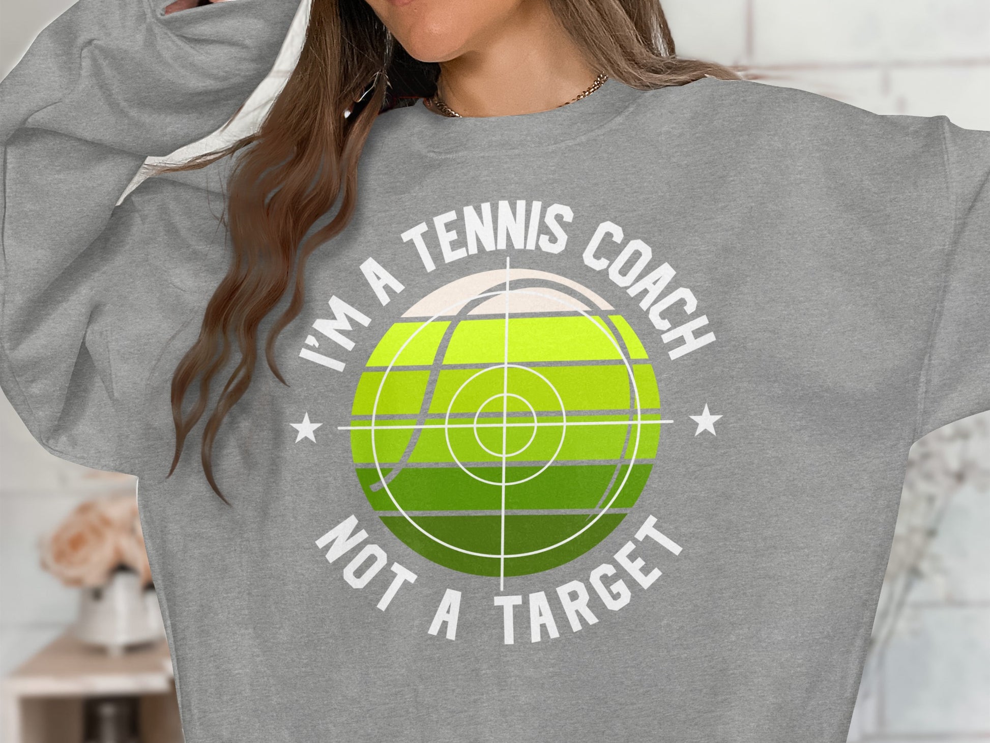 Gray sweatshirt with "I'm a tennis coach, not a target" text and tennis ball graphic.