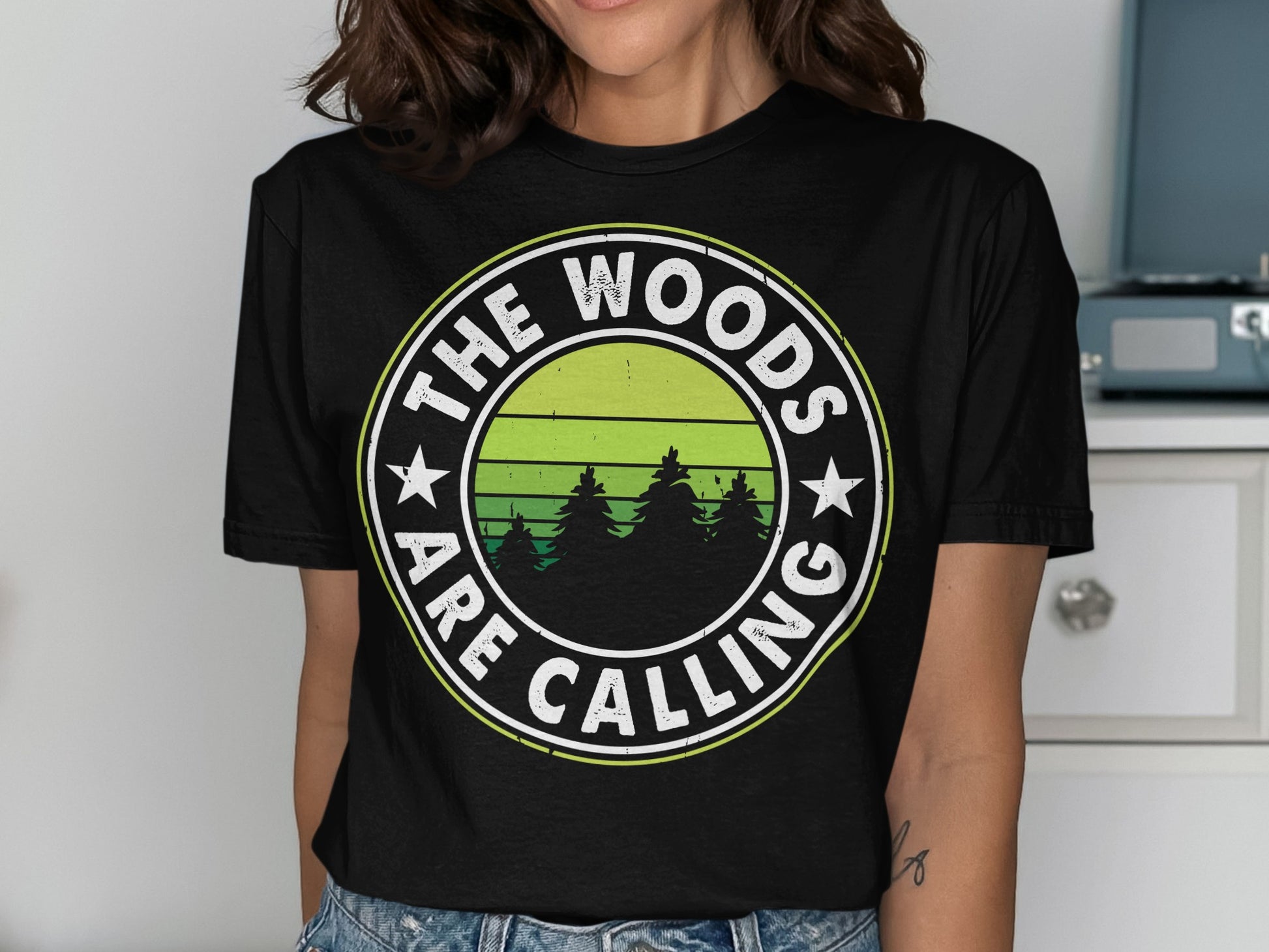 Black t-shirt with circular graphic, "The woods are calling," and green trees.