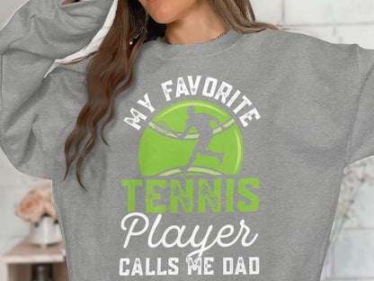 Gray sweatshirt with text "MY FAVORITE TENNIS PLAYER CALLS ME DAD" and a tennis ball graphic.